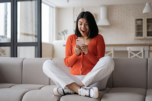 Young emotional woman holding smartphone playing mobile game celebration success at home. Excited African American hipster using mobile phone shopping online with big sales sitting on sofa