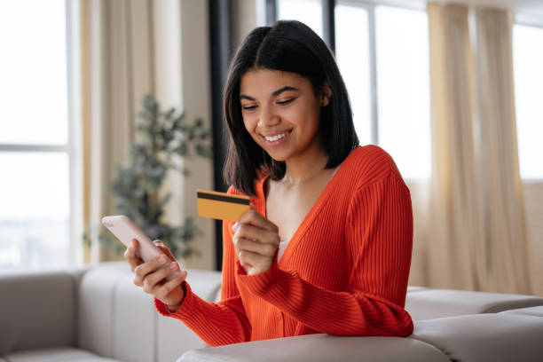Smiling African American woman holding credit card using mobile phone shopping online, check card balance. Mobile banking. Beautiful female ordering food online stock photo