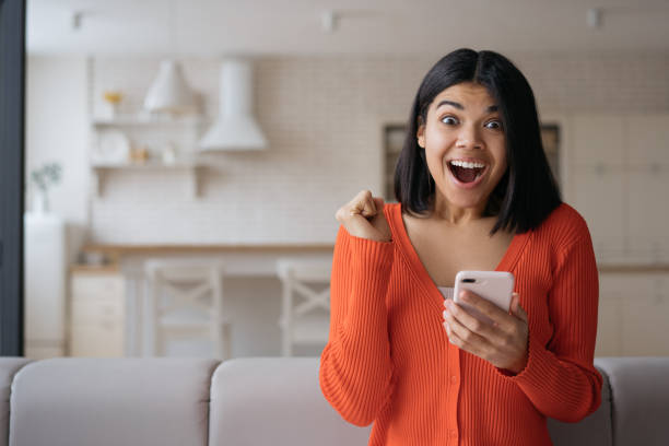 Excited African American using mobile phone shopping online with big sales. Happy female win online lottery celebration success sitting at home. Emotional hipster playing mobile game, copy space stock photo