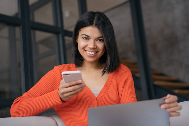 Confident businesswoman using laptop computer holding mobile phone sitting in modern office. Smiling African American freelancer working online from home. Successful business stock photo