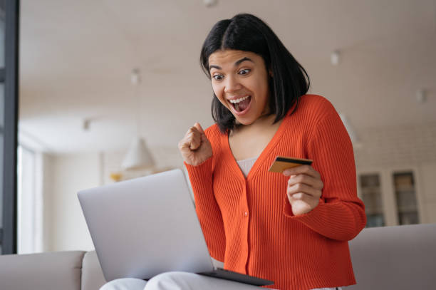 Overjoyed African American woman holding credit card using laptop computer shopping online, check card balance. Excited female ordering food online. Happy freelancer receive payment working from home stock photo