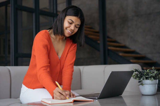 istock Confident businesswoman using laptop computer taking notes working in modern office. Smiling African American student studying, online education concept 1387323642