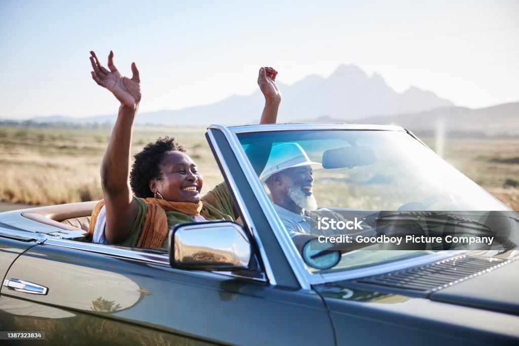 Smiling woman having fun during a scenic road trip with her husband in summer Mature African woman smiling with her hands raised in the air during a scenic road trip with her husband in their convertible Road Trip Stock Photo