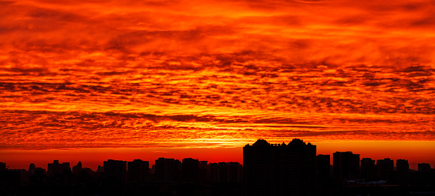 A bloody dawn and a panorama of the silhouettes of the buildings of the night city against the backdrop of a bright red sunrise in the early morning. Black and red landscape of night Kyiv.