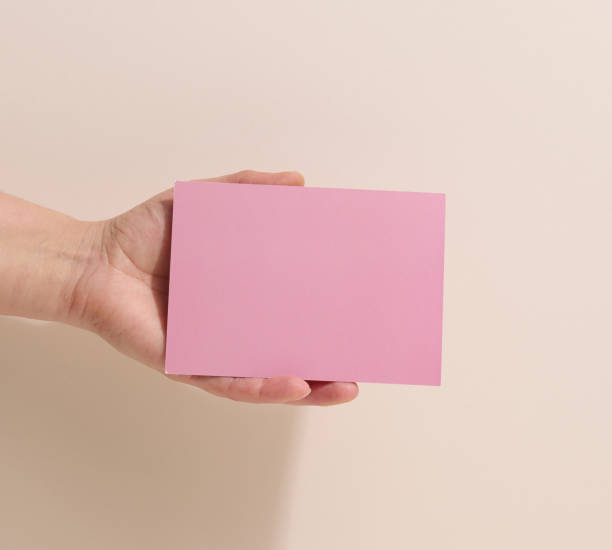 istock Female hand holding empty pink paper on a beige background. Copy paste image or text, close up 1387338459