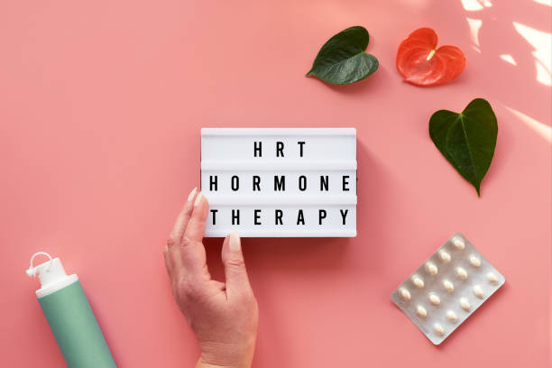 Text HRT Replacement Therapy on light box in hand. Menopause, hormone therapy concept. Pink background with exotic leaves, flowers, pills, estrogene gel stock photo
