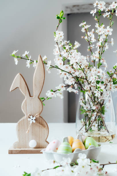 lapin de pâques en bois avec branche d’arbre en fleurs avec des fleurs dans le vase et des œufs de pâques colorés dans un récipient sur la table blanche. bonne carte verticale de printemps de pâques. mise au point sélective. espace de copie. - figurine easter egg easter holiday photos et images de collection