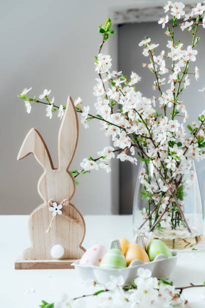 istock Wooden Easter bunny with blooming tree branch with flowers in the vase and colored easter eggs in container on the white table. Happy Easter spring vertical card. Selective focus. Copy space. 1387339988