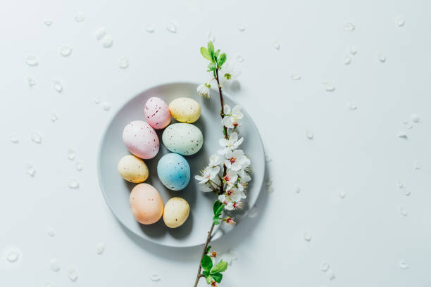 istock Top view Colored Easter eggs and blooming tree branch on the gray plate with flower petals on the white table. Happy Easter and springtime card. Minimalism and simplicity. Selective focus. Copy space. 1387339989