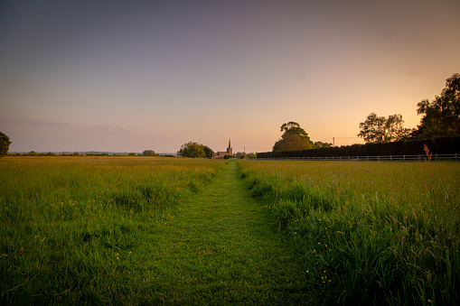 A footpath in a green field stretches towards a church at sunset.