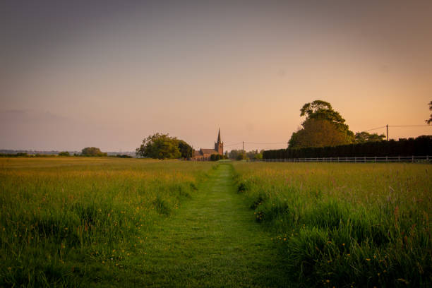 A footpath in a green field stretches towards a church at sunset.
