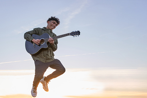 Happy Latin teenager jumping with guitar over the blue sky at sunset.