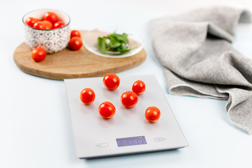 Red cherry tomatoes on gray digital kitchen scales. Near bowl with tomatoes, plate with fresh green basil on wooden board and towel. Close up, flat lay. Kitchen equipment concept. Weighing products.