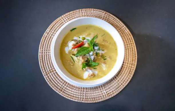 Photo of Flatlay Top Table of Green Curry with Chicken garnished on top with pepper, basil was served in white bowl on wicker placemat cream color. Clipping Path.