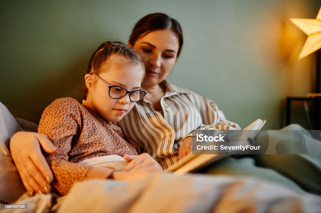 Mother and Daghter Reading Book at Night Portrait of little girl with down syndrome reading book at bedtime and relaxing with caring mother embracing her Child Stock Photo