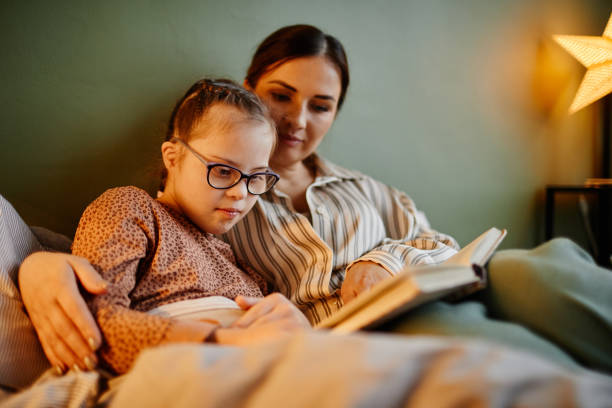 Mother and Daghter Reading Book at Night Portrait of little girl with down syndrome reading book at bedtime and relaxing with caring mother embracing her developmental disability diversity stock pictures, royalty-free photos & images