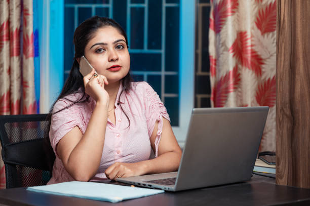 istock Smiling female Indian employee multitask working on laptop computer in office talking over phone. 1387415835