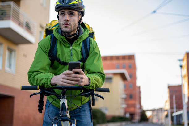 handsome young man using bicylce to deliver boxes, documents and food at home, male courier using technology of smart devices to find address and sending work report