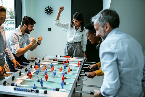 Coworkers playing table football on break from work. Diverse group of colleagues having fun entertainment enjoyment. Business people having great time together.Colleagues playing table football in modern office