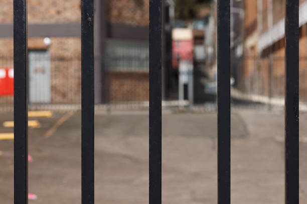 Black iron bars with car park and brick wall beyond Black metal fence. prison lockdown stock pictures, royalty-free photos & images