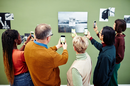 Group of multi-ethnic visitors scanning QR code to get more information about black and white photo on wall at exhibition in museum