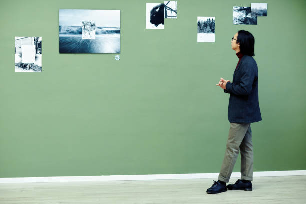 Young Asian man with long hair wearing eyeglasses spending time in modern art gallery looking at black and white photos on green wall at exhibition