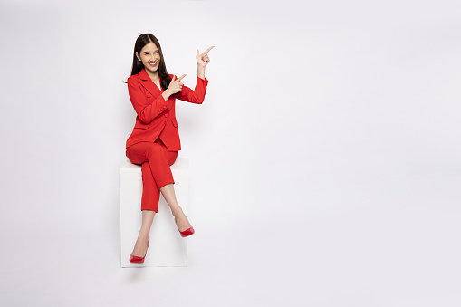 Young Asian businesswoman in red suit sitting and pointing to empty copy space isolated on white background