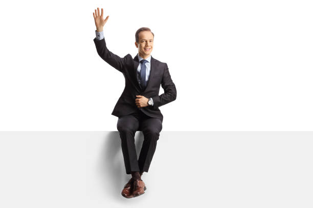 istock Professional man in suit and tie sitting on a blank panel and waving 1387428445