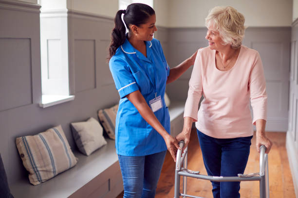 ältere frau zu hause mit gehhilfe, die von einer pflegerin in uniform unterstützt wird - physical and occupational therapists stock-fotos und bilder