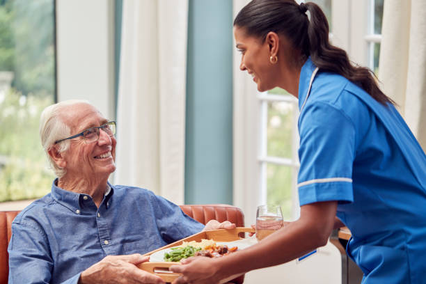 Female Care Worker In Uniform Bringing Meal On Tray To Senior Man Sitting In Lounge At Home stock photo