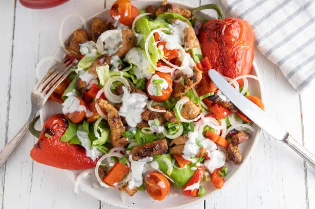 Delicious and healthy salad dish with pan fried turkey meat, roasted bell peppers and tomatoes, onions, lettuce and a low fat yogurt, garlic, herb dressing. Served on a white plate on white table background. Overhead view and closeup
