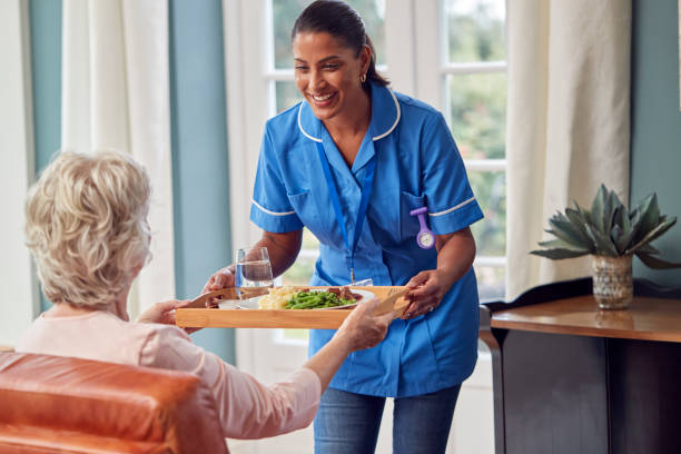 Female Care Worker In Uniform Bringing Meal On Tray To Senior Woman Sitting In Lounge At Home stock photo