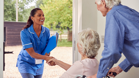 Pareja de ancianos con mujer en silla de ruedas saludando a enfermera o cuidadora haciendo visita domiciliaria en la puerta photo