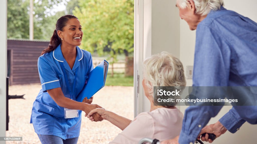 Senior Couple With Woman In Wheelchair Greeting Nurse Or Care Worker Making Home Visit At Door Home Caregiver Stock Photo