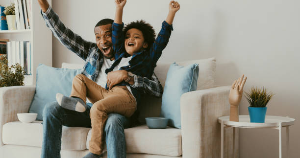 père et fils regardant la télévision à la maison. encourageant. - child celebration cheering victory photos et images de collection