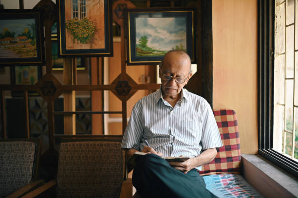 Senior man sitting next to window and writing in a diary. He is an Author and a Poet.