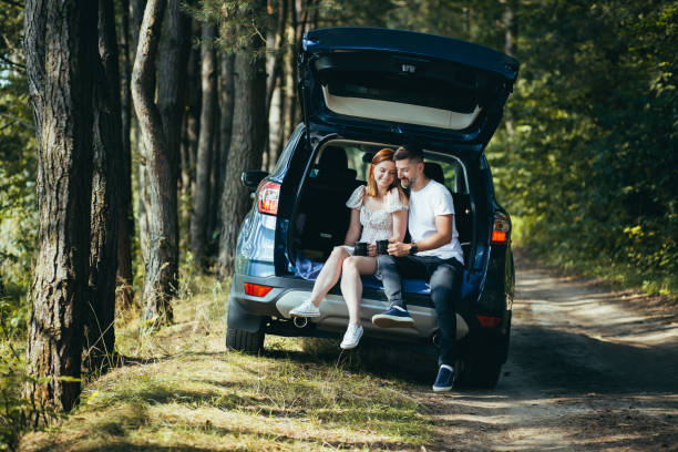 giovane coppia, uomo e donna, abbracciati insieme durante un picnic, seduti nel bagagliaio di un'auto nel bosco, felici insieme - car family picnic vacations foto e immagini stock