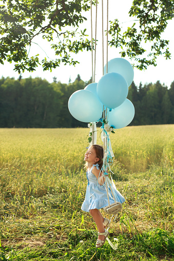 A girl in an elegant dress with balloons swinging on a swing and laughing loudly with happiness. The child is celebrating his fourth birthday. Sweet baby enjoys the moment