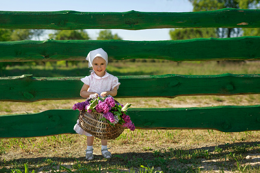 A girl in a kerchief and with a basket in her hands full of purple lilac flowers. The child collected a bouquet of spring flowers in the flowering season. Copy space