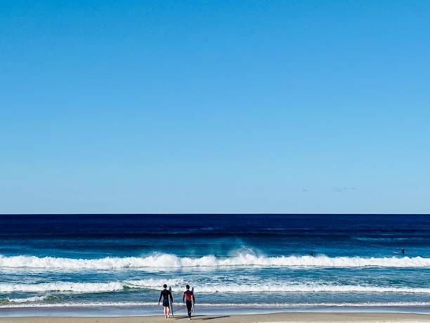 Horizontal seascape of outlined unidentifiable surfers with surfboards looking out to turquoise breaking waves in calm ocean waters under a clear blue sky at Brunswick Heads near Beach Byron Bay NSW Australia