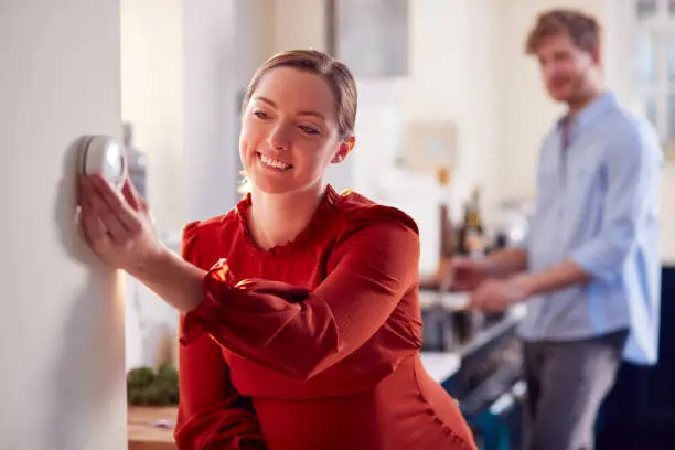 Photo of Pregnant Couple With Woman With Prosthetic Arm Turning Down Heating Control In Kitchen Together