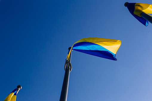 Color image depicting the national flag of Ukraine raised on flagpoles and proudly flying in the wind against a clear blue sky.