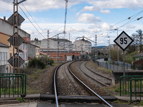 Train tracks at the entrance to Monforte de Lemos station from a level crossing with barriers for pedestrians and a maximum speed signal on the side of the track