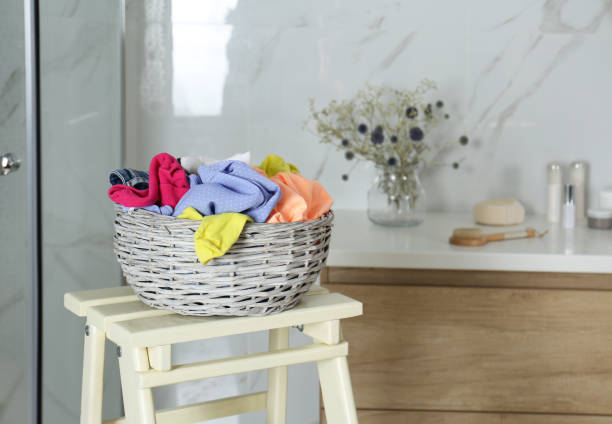 istock Wicker laundry basket with different clothes on wooden stool in bathroom. Space for text 1387463218