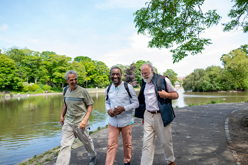 Three senior men exploring leazes park and enjoying a day out together in Newcastle Upon Tyne, England. They are walking on a footpath next to a lake while they walk, talk and smile.