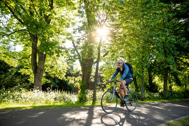 Saving the Planet and Keeping Healthy A senior man wearing sunglasses, riding his bike on a footpath through a public park in Newcastle upon Tyne, England. He is smiling while the sun shines down on him. wide shot stock pictures, royalty-free photos & images