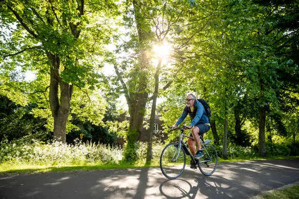 A senior man wearing sunglasses, riding his bike on a footpath through a public park in Newcastle upon Tyne, England. He is smiling while the sun shines down on him.