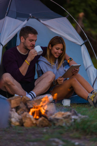 istock Young couple drinking coffee, enjoying the mountain view and checking the tablet with bonfire in the sunset 1387464586