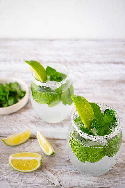 Two glasses with summer lemonade with lime and mint stand on a wooden table. Nearby lie three slices of lime and there is a bowl with mint. The edges of the glasses are coated with sugar. Vertically Two glasses with summer lemonade with lime and mint stand on a wooden table. Nearby lie three slices of lime and there is a bowl with mint. The edges of the glasses are coated with sugar. Vertically soda water glass lemon stock pictures, royalty-free photos & images
