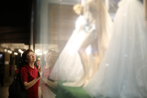 An Asian woman is admiring bridal gown at bridal boutique display.